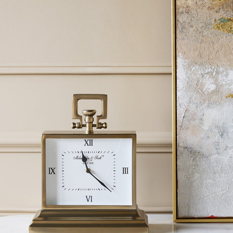 Libra Interiors brushed brass rectangular mantel clock with white Roman numeral dial, loop handle and stepped base, styled beside a gold framed artwork.