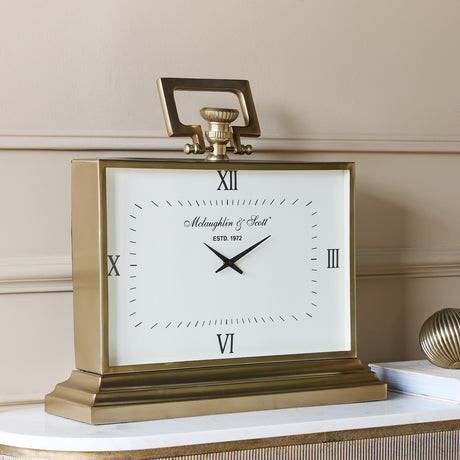 Libra Interiors brushed brass rectangular mantel clock with white Roman numeral dial, looped handle and stepped base on a marble console.