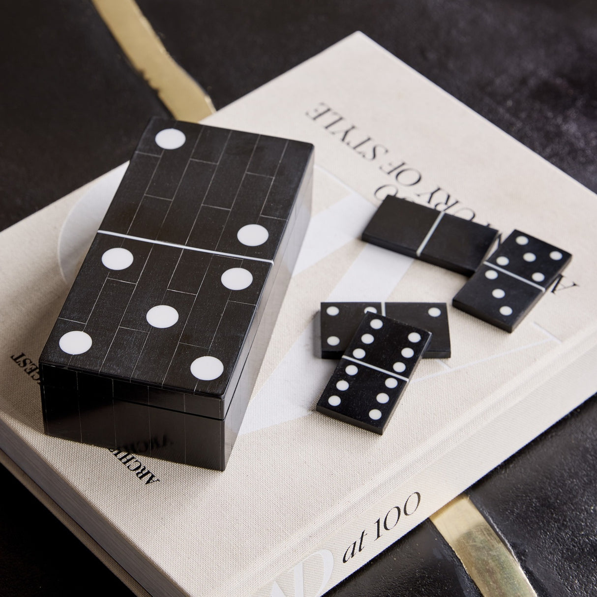 Libra Interiors Resin Inlay Black Dominoes Game shaped as an oversized domino tile with white spots, shown on a book with loose pieces beside it.