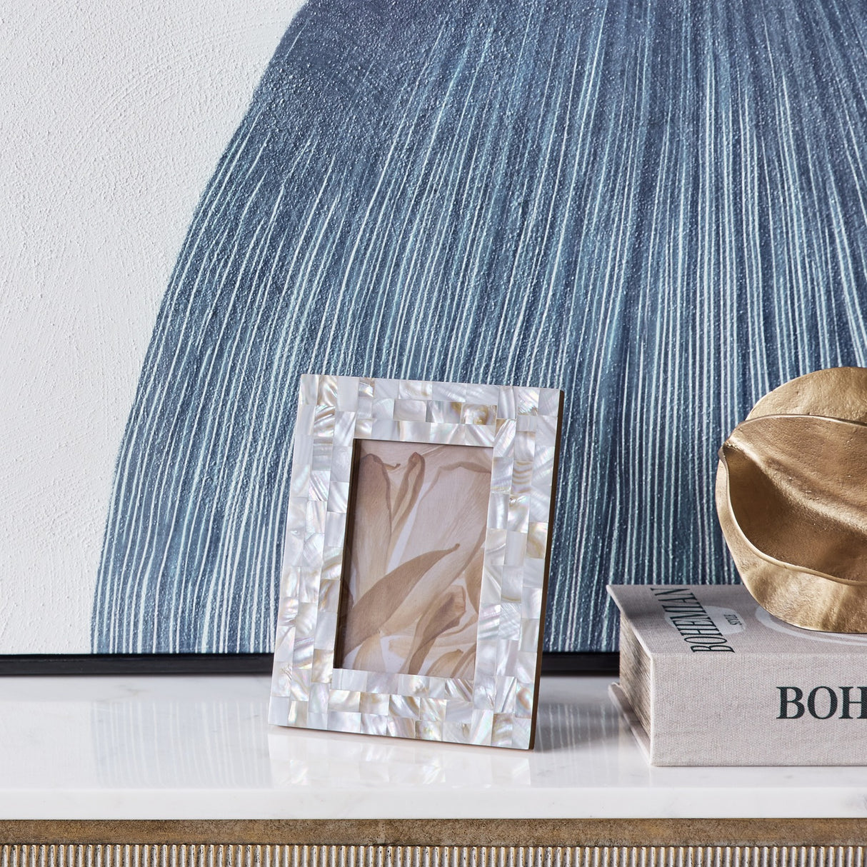 Libra Interiors Mother of Pearl Mosaic 4x6 photo frame in ivory on a marble console, styled beside a blue textured artwork, book and gold leaf decor.