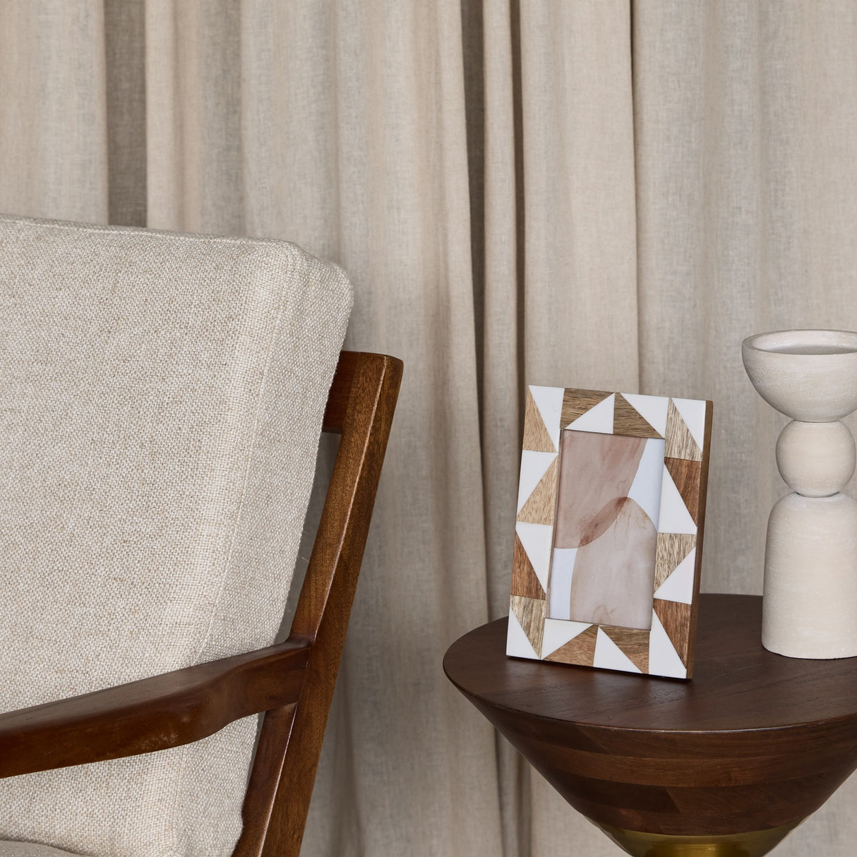 Libra Interiors Geometric Wood and Resin Photo Frame in brown and cream triangular inlay, on a walnut side table beside a ceramic vase and linen curtains.
