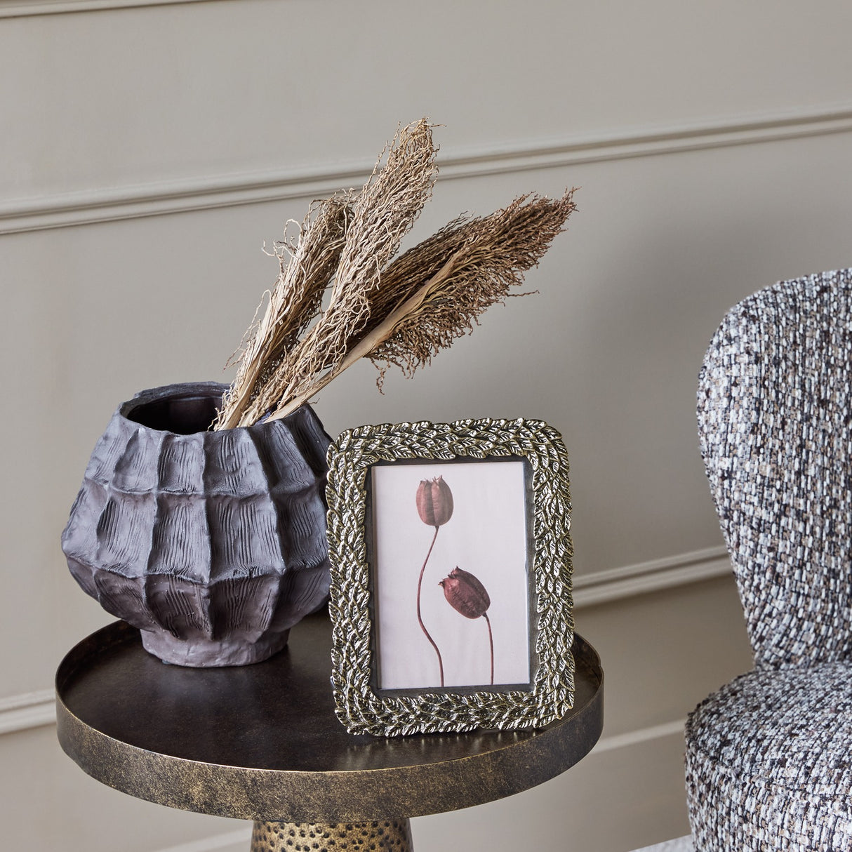 Libra Interiors Antiqued Leaf gold aluminium 5x7 photo frame on a bronze side table beside a textured dark vase with dried grasses and a woven chair.