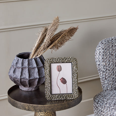 Libra Interiors Antiqued Leaf gold aluminium 5x7 photo frame on a bronze side table beside a textured dark vase with dried grasses and a woven chair.