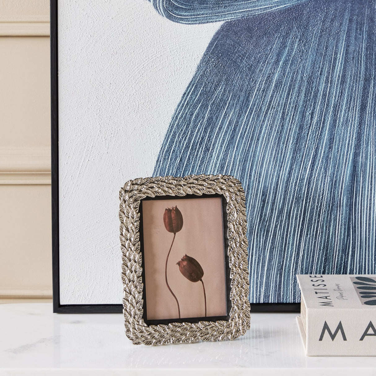 Libra Interiors Antiqued Leaf Aluminium Photo Frame in silver, displayed on a marble surface beside blue artwork and Matisse books in a living room setting.