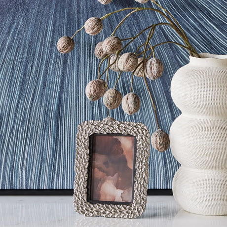 Libra Interiors Antiqued Leaf aluminium photo frame in silver, displayed beside a textured cream vase and blue linear artwork on a marble surface.