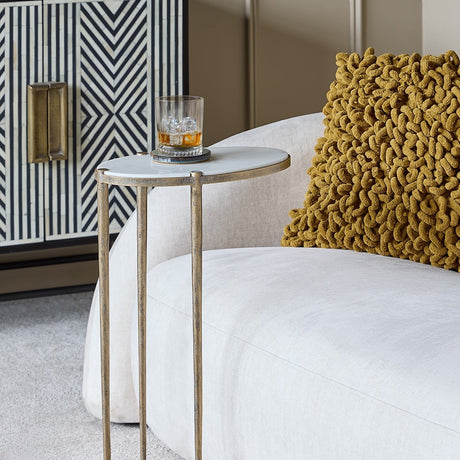 Libra Interiors Marble Top Oval Side Table in gold holding a whisky glass beside a cream armchair with mustard cushion and striped cabinet.