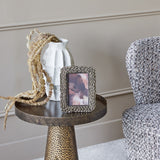 Libra Interiors Round Marble Side Table in grey with aged silver metal base, styled beside a tweed armchair with a white vase and dried grasses on top.