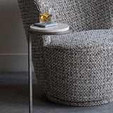 Libra Interiors Round Marble Side Table in grey, tucked beside a textured bouclé armchair with a whisky glass on marble coasters atop its round marble surface.