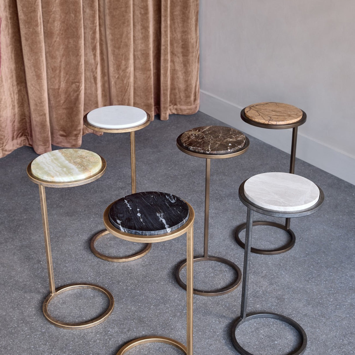 Libra Interiors Round Marble Side Tables grouped on grey flooring, showing varied stone tops in white, green, black, brown and grey with metal frames.