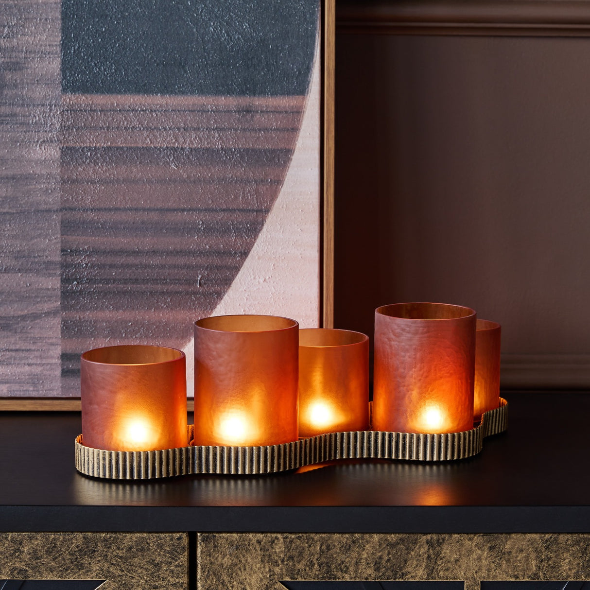Libra Interiors Hammered Glass 5 Light Amber Lantern lit on a dark console, five frosted amber votives on a ribbed aged gold base beside artwork.