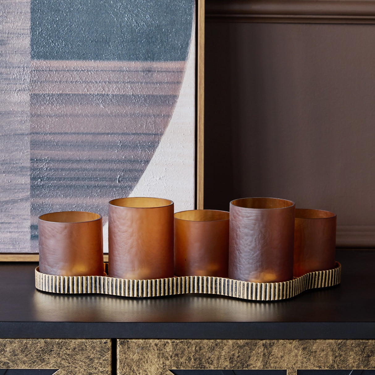 Libra Interiors Hammered Glass 5 Light Amber Lantern with amber votives at stepped heights on a ribbed aged gold base, styled on a dark console.