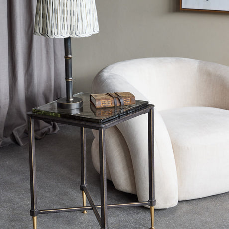 Libra Interiors Textured Glass Top Iron Side Table in black with brass-tone feet, holding a lamp and leather journal beside a cream armchair.