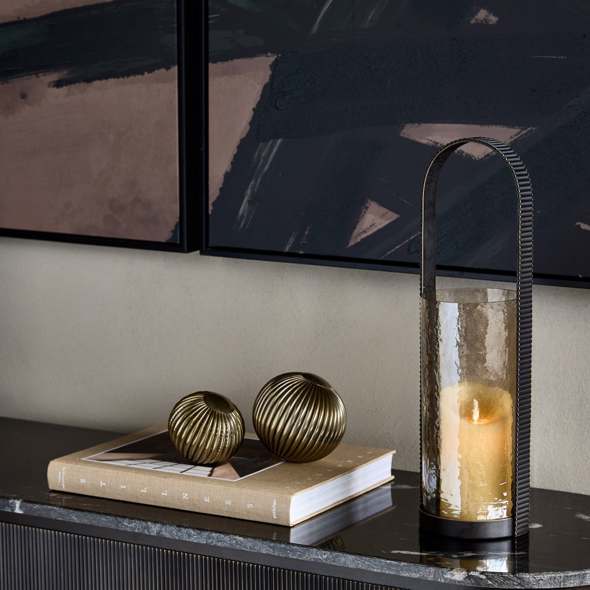 Libra Interiors hammered glass hurricane lantern with copper-brown cylinder and arched corrugated iron handle, styled on a marble console with brass decor.