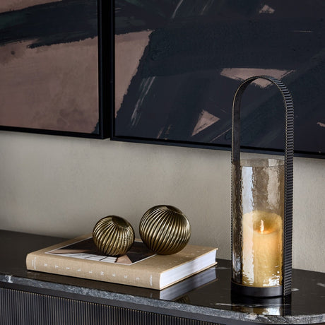 Libra Interiors hammered glass hurricane lantern with copper-brown cylinder and arched corrugated iron handle, styled on a marble console with brass decor.
