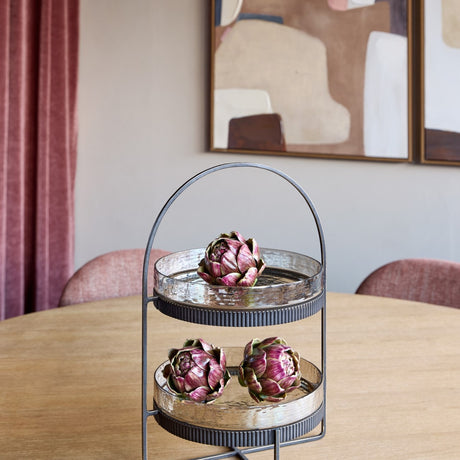 Libra Interiors Hammered Glass Two Tier Cake Stand in bronze with arched handle, displayed with artichokes on a dining table beside pink curtains.