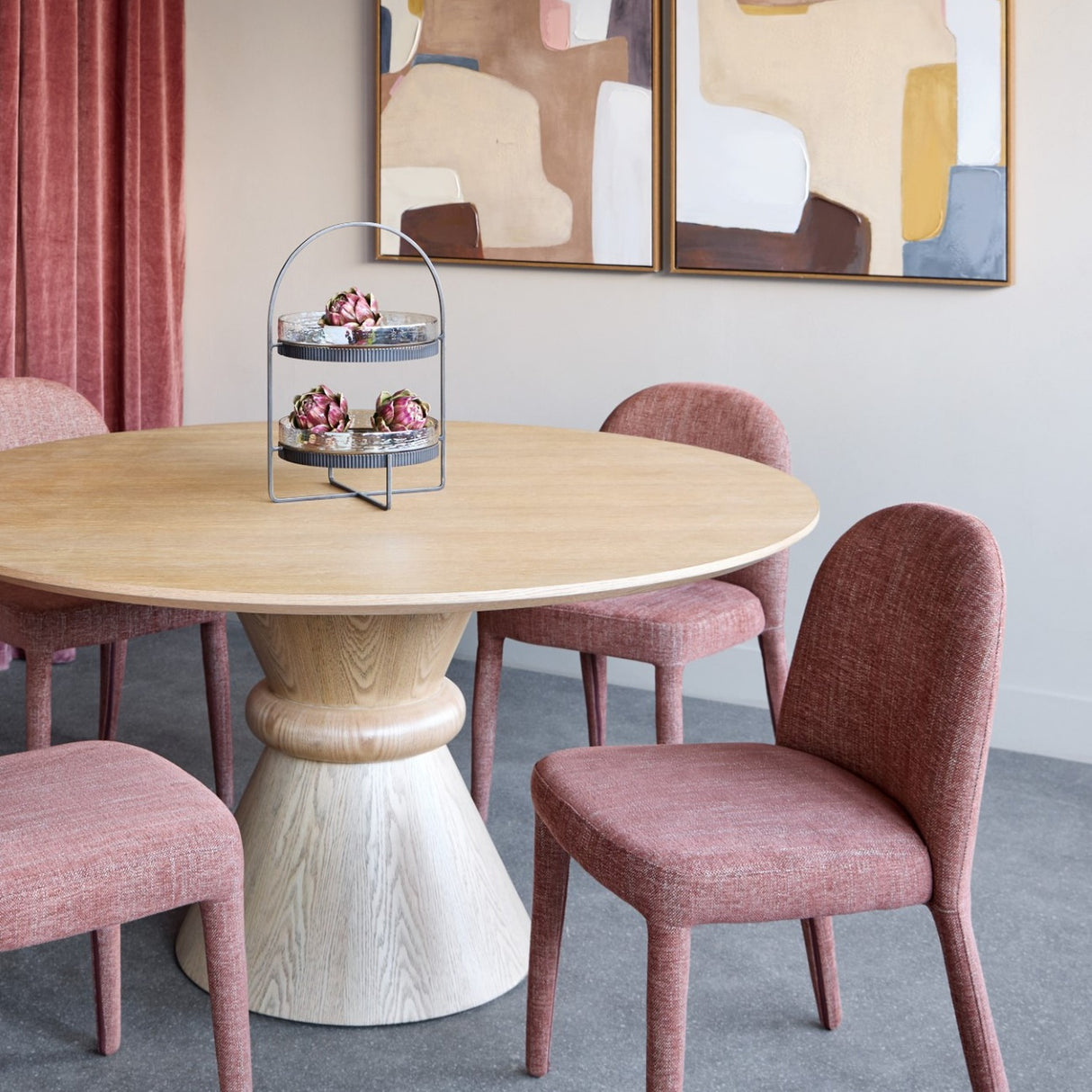 Libra Interiors Hammered Glass Two Tier Cake Stand in bronze holding artichokes on a round oak dining table with pink chairs and abstract art.