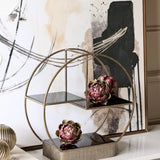 Libra Interiors Aged Gold tiered circular cake stand with antiqued iron frame and brown tinted glass shelves, styled with artichokes beside abstract wall art.