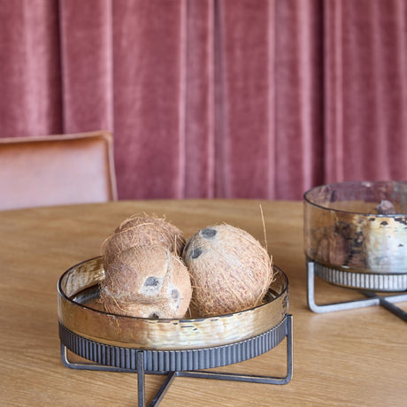 Libra Interiors Textured Glass and Iron Cake Stand in bronze holding coconuts on a wooden table, with a matching bowl and pink velvet curtains behind.
