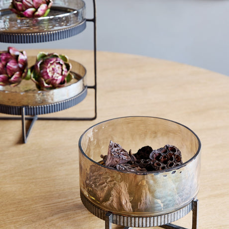 Libra Interiors Hammered Glass & Iron Cake Stand in bronze holding dried lotus pods, styled beside a two-tier stand with artichokes on a wooden table.