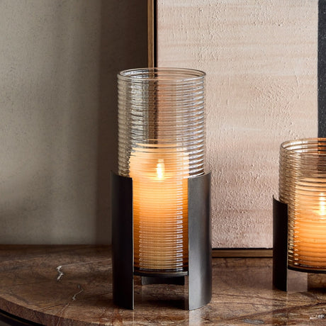 Libra Interiors Ribbed Glass Bronzed Iron Hurricane Lantern in brown, lit with a pillar candle on a marble table beside a smaller matching lantern.