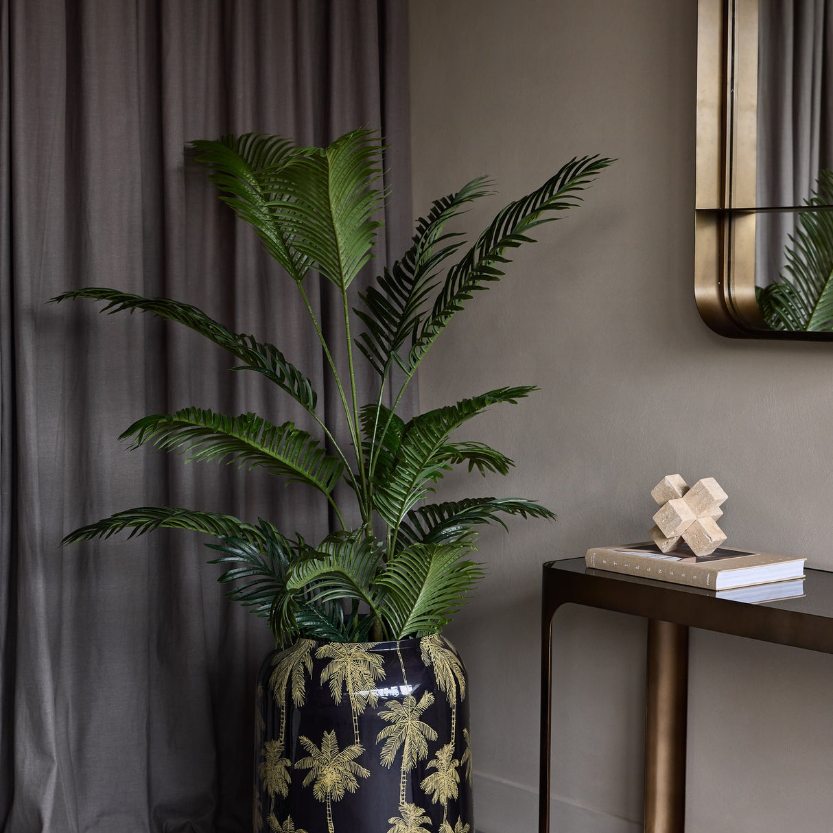 Libra Interiors Gold Palm Enamel Iron Vase in black holds tall green palm foliage beside a bronze console with mirror and grey curtain in a styled hallway.