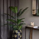 Libra Interiors Gold Palm Enamel Iron Vase in black holds tall green palm foliage beside a bronze console with mirror and grey curtain in a styled hallway.