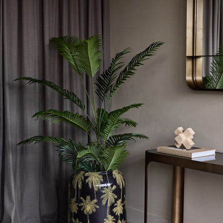 Libra Interiors Gold Palm Enamel Iron Vase in black holds tall green palm foliage beside a bronze console with mirror and grey curtain in a styled hallway.
