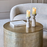 Libra Interiors Drum Storage Coffee Table in champagne gold aluminium, styled with two candle holders and a book beside a cream bouclé armchair.