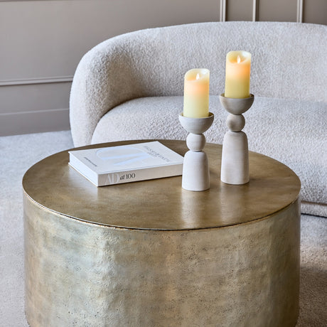 Libra Interiors Drum Storage Coffee Table in champagne gold aluminium, styled with two candle holders and a book beside a cream bouclé armchair.