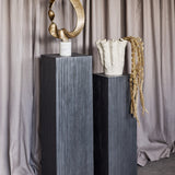 Libra Interiors Grooved Wood Black Display Plinths in matt black mango wood, styled with a gold sculpture and white ceramic vase against draped linen.