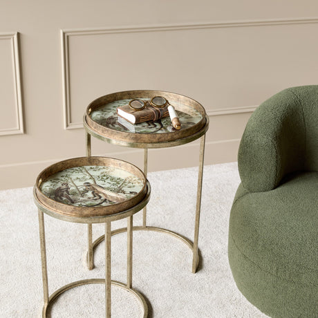 Libra Interiors Removable Tray Top Round Nesting Side Tables in antique gold with monkey-scene glass trays, styled beside a green bouclé armchair.
