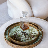 Libra Interiors Round Glass Monkey Jungle serving trays in cream and gold with antiqued frames, holding a crystal decanter on a pale upholstered seat.