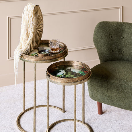 Libra Interiors gold nesting side tables with round removable trays showing tropical birds artwork, styled beside a green boucle armchair in a living room.