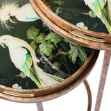 Close-up of Libra Interiors Removable Tray Top Nesting Side Tables in gold, showing round trays with tropical cockatoo and foliage prints under glass.