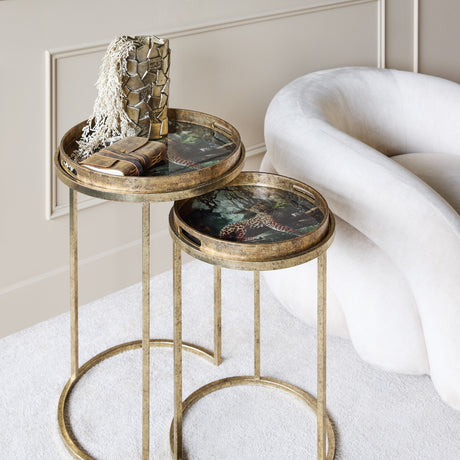 Libra Interiors Antiqued Gold Leopard nesting side tables with round tray tops and slender metal legs, styled beside a cream armchair in a living room.
