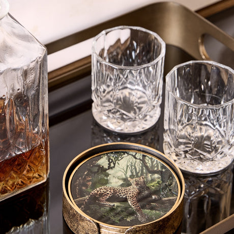Libra Interiors Jungle Print round acrylic coaster with leopard illustration and antiqued gold rim, styled on a bar tray beside crystal glasses and a decanter.