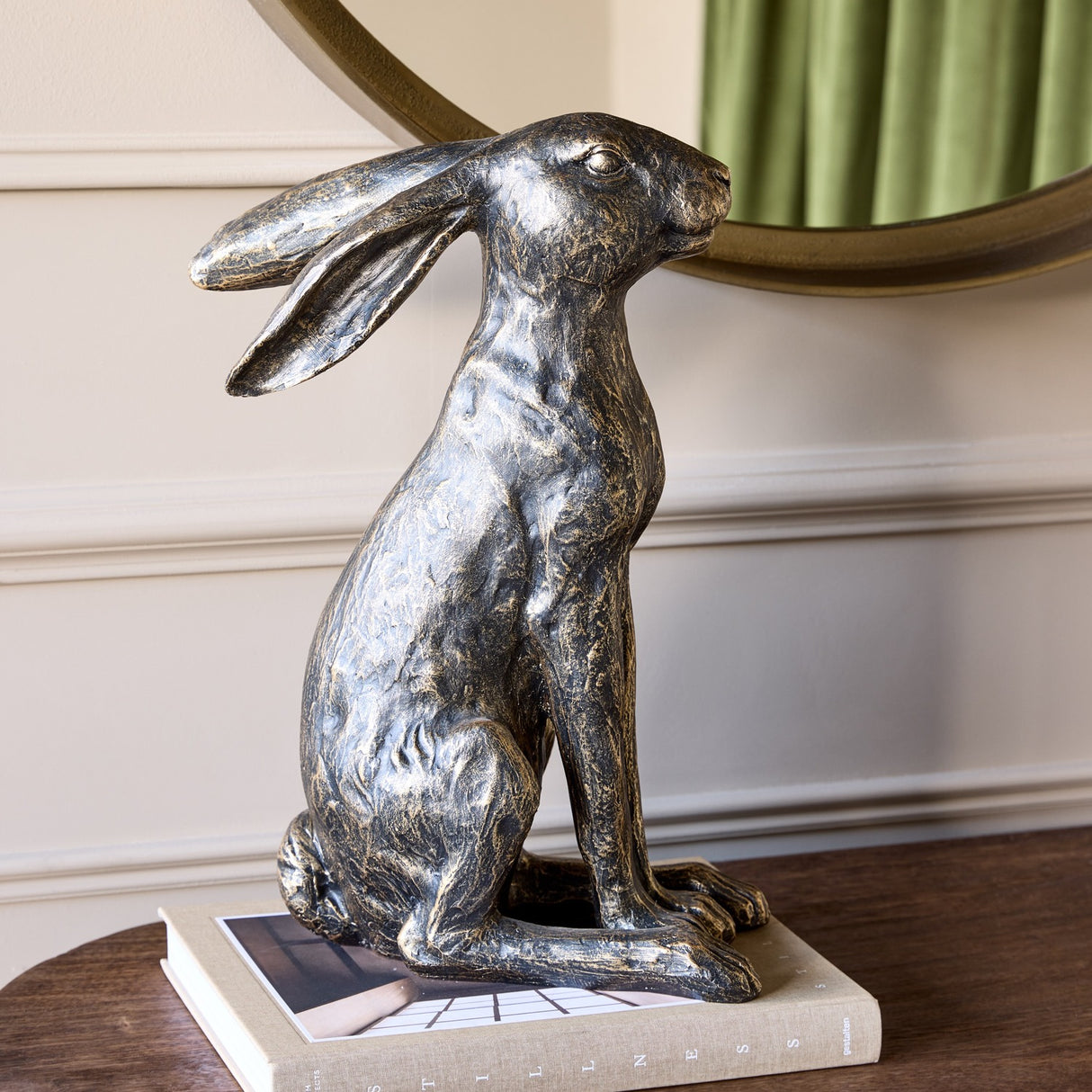 Libra Interiors Antique Bronze Sitting Hare Sculpture with textured finish and long ears, displayed on a book atop a wooden console beneath a round mirror.