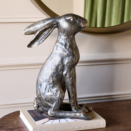 Libra Interiors Antique Bronze Sitting Hare Sculpture with textured finish and long ears, displayed on a book atop a wooden console beneath a round mirror.