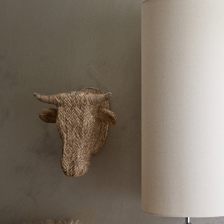 Libra Interiors Braided Resin Cow Head Wall Art in brown, mounted on a taupe wall beside a tall cream cylindrical lamp shade in a rustic interior.