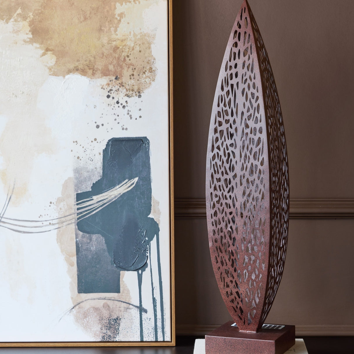 Libra Interiors Solar Cut-Out Iron Leaf Sculpture in bronze, tall pierced leaf form on square base, displayed beside an abstract framed artwork.