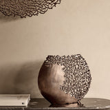 Libra Interiors Coral Cut-Out Aluminium Ellipse Vase in bronze on a marble console, with coral-inspired lattice detailing beside a book against a neutral wall.