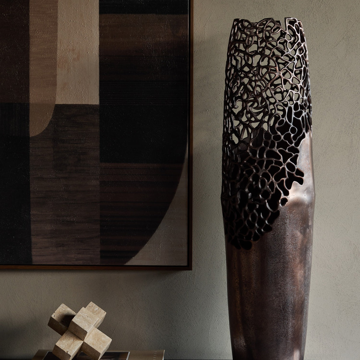 Libra Interiors Coral Cut-Out bronze aluminium floor vase with smooth base and pierced openwork top, styled beside abstract art and a travertine sculpture.