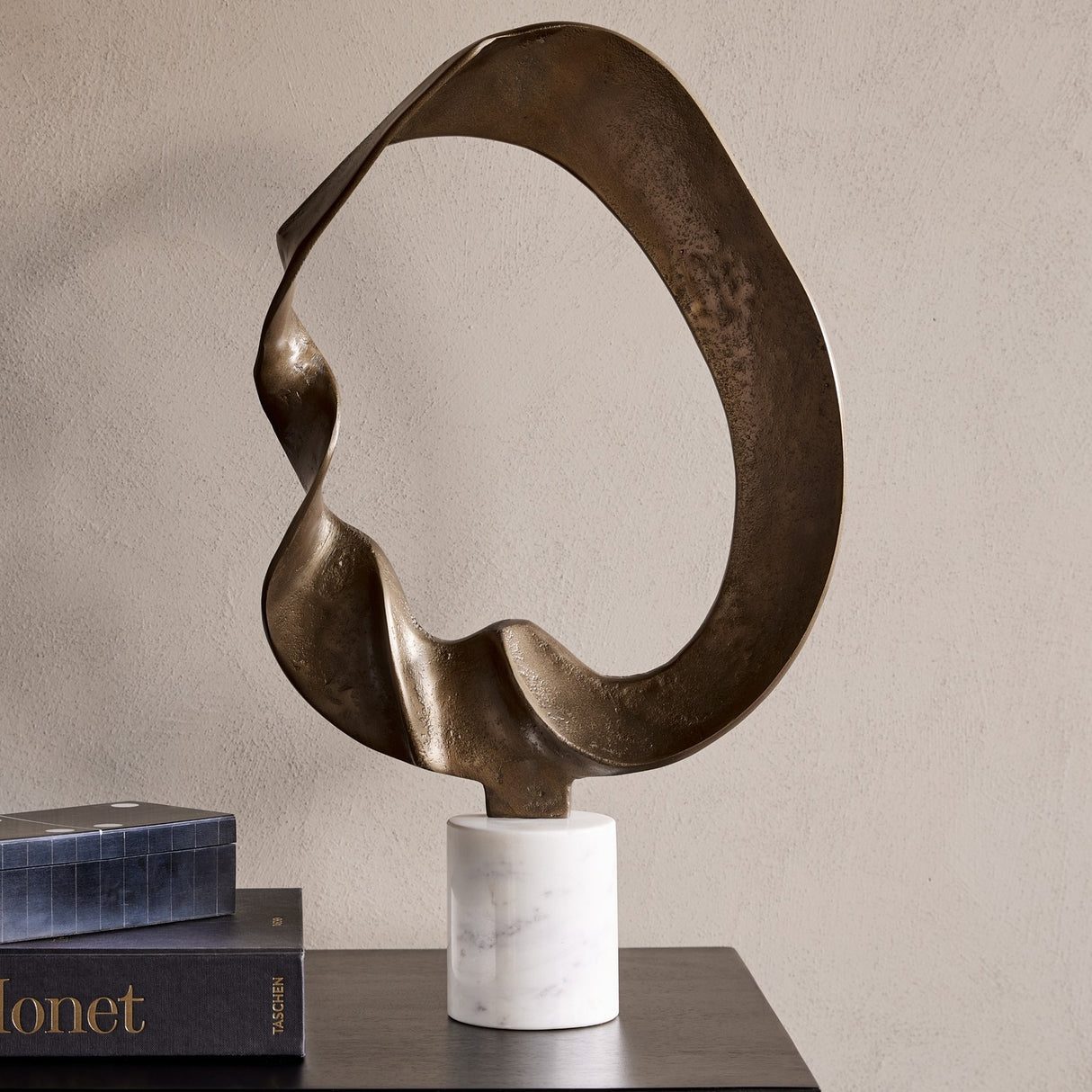 Libra Interiors Abstract Aluminium Ring Sculpture in antique gold with a looping textured form on a white marble base, styled beside books on a console.