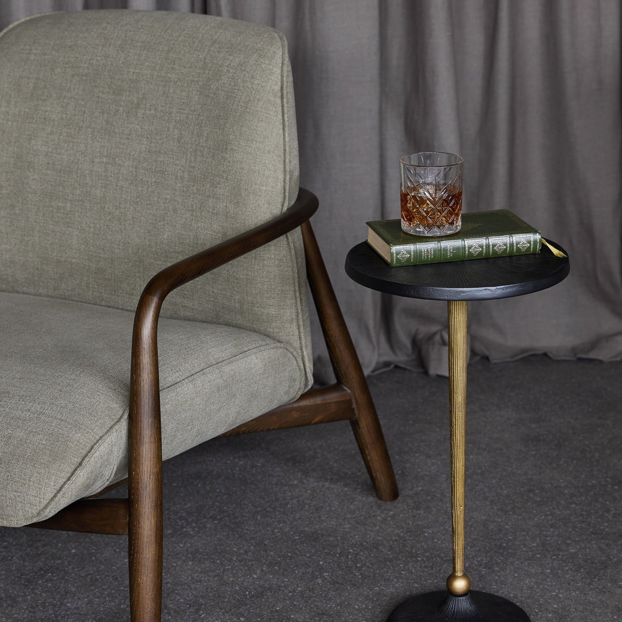 Libra Interiors Grooved Sunburst round side table with blackish bronze top and matt gold stem, styled beside an armchair with a book and whisky glass.