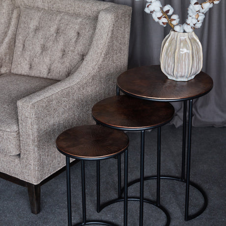 Libra Interiors Radial Etched round nesting side tables in bronze with black frames, staggered beside a grey armchair with a cotton stem vase on top.