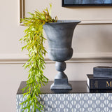 Libra Interiors Stonewash Grey Aluminium Urn Vase with flared lip and spherical stem on a console, holding trailing green foliage beside stacked books.