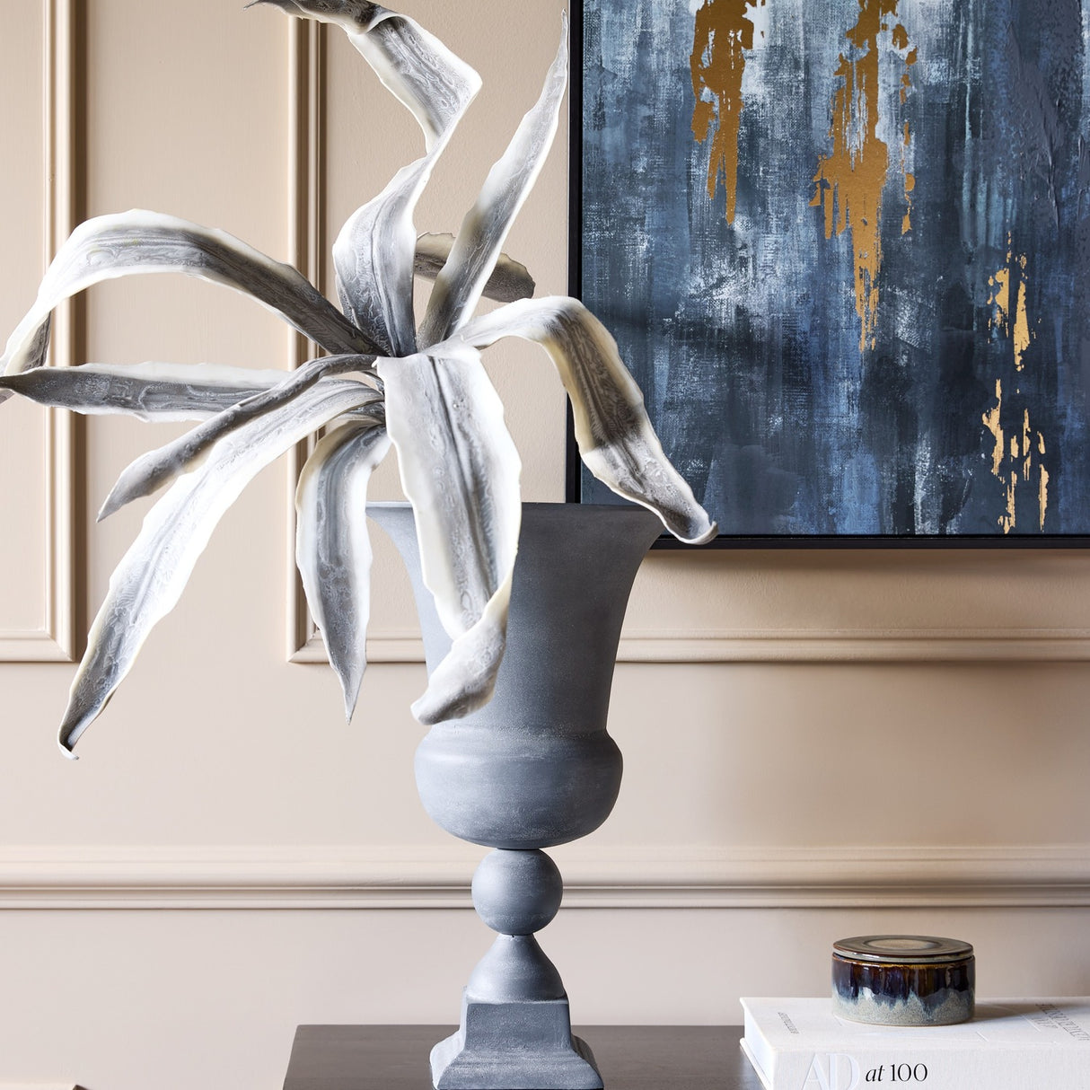 Libra Interiors Stonewash grey aluminium urn vase with flared opening and square plinth, holding tall grey foliage beside abstract blue and gold artwork.