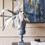 Libra Interiors Stonewash grey aluminium urn vase with flared opening and square plinth, holding tall grey foliage beside abstract blue and gold artwork.