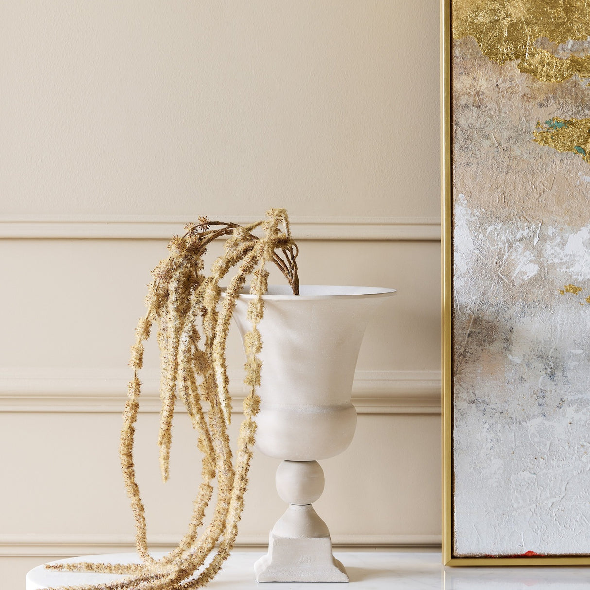 Libra Interiors Stonewash Aluminium Urn Vase in cream with flared rim and stepped plinth, holding dried stems beside a gold-toned abstract artwork.