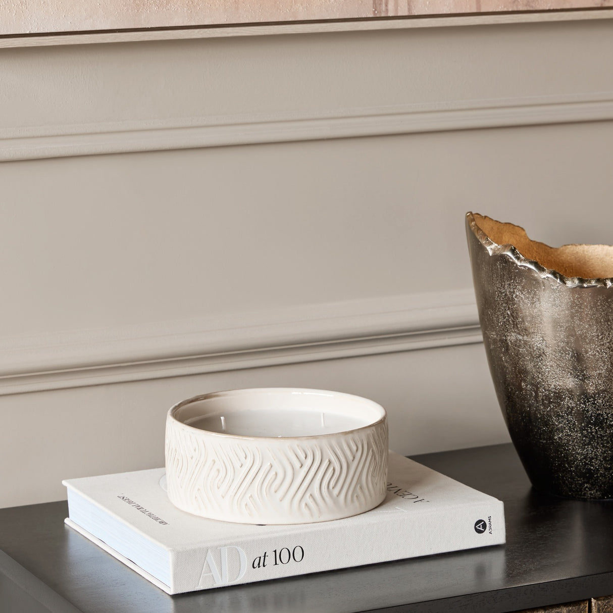 Libra Interiors Embossed Glazed Ceramic Bowl Candle in ivory with a flowing geometric relief, resting on a white AD at 100 book beside a dark metallic vase.